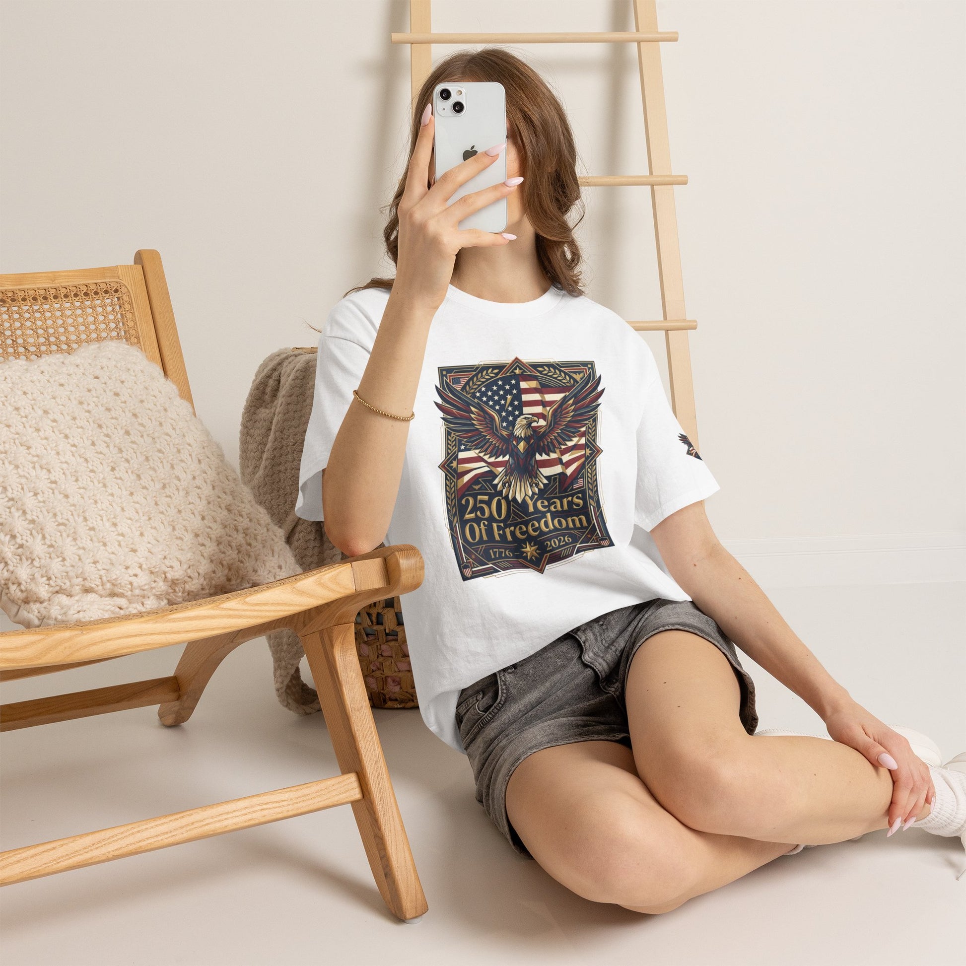 250 Years of Freedom Eagle T-Shirt, Woman sitting on the floor holding a phone, wearing a white t-shirt with a graphic design.