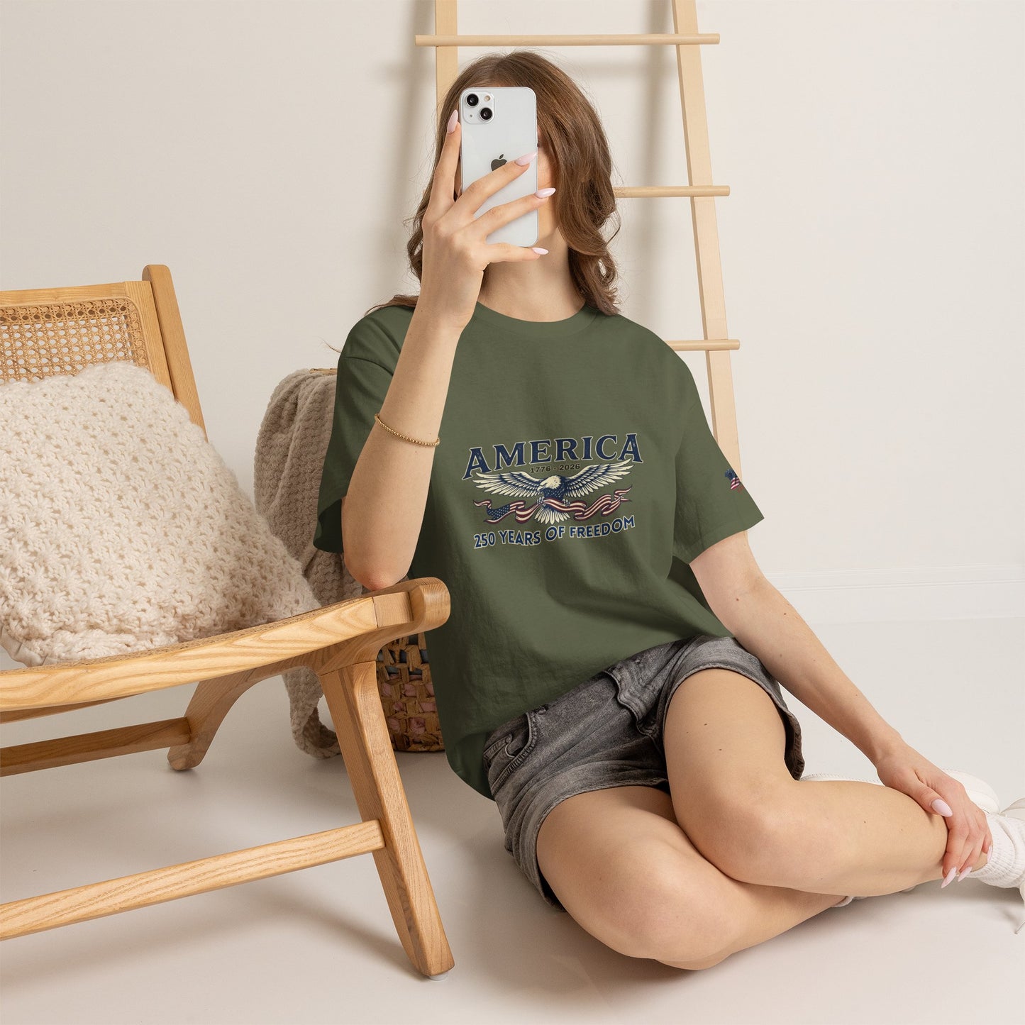 America 250 Years of Freedom T-Shirt — USA 250th ANNIVERSARY T-shirt, Person wearing a green t-shirt with a graphic design, sitting on the floor in a room with a wooden chair and ladder.
