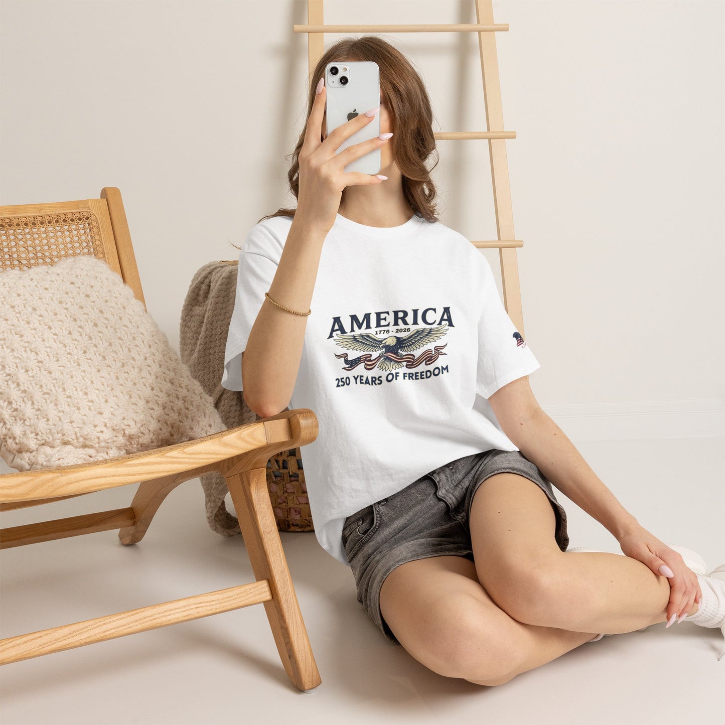 America 250 Years of Freedom T-Shirt — USA 250th ANNIVERSARY T-shirt, Person wearing a white t-shirt with 'AMERICA' text, sitting on the floor with a phone.